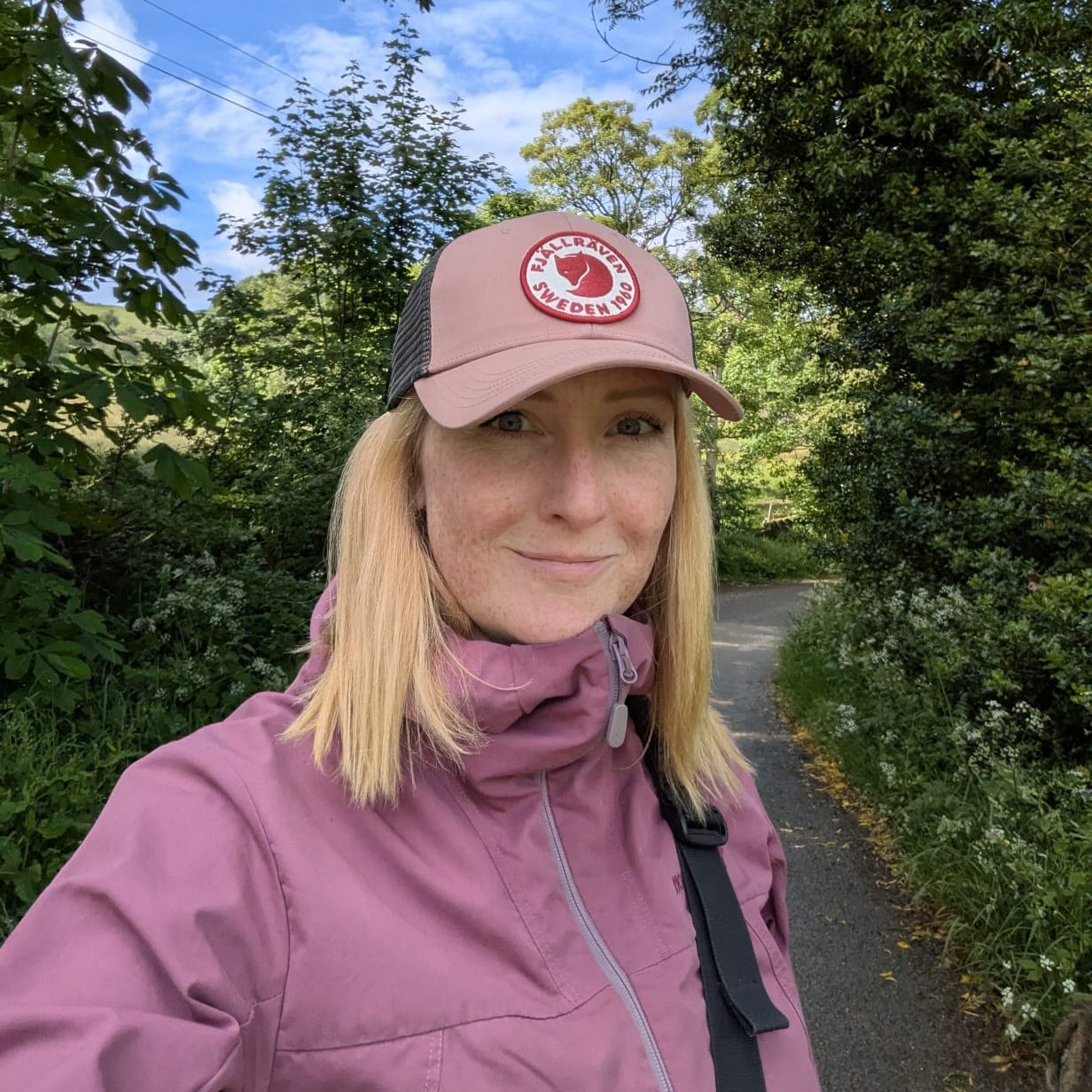 Jo Kelly, founder of White Peak Pets in a pink jacket and cap stands on a path surrounded by greenery.