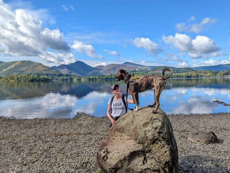 Jo Kelly stands by a lake, training Rupert the dog standing on a rock, surrounded by mountains and clouds.